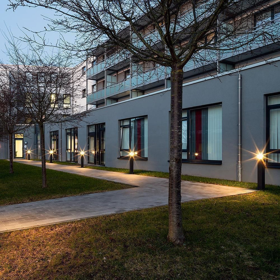 Allée bordée de plusieurs bornes lumineuses au crépuscule, arbres et bâtiment.