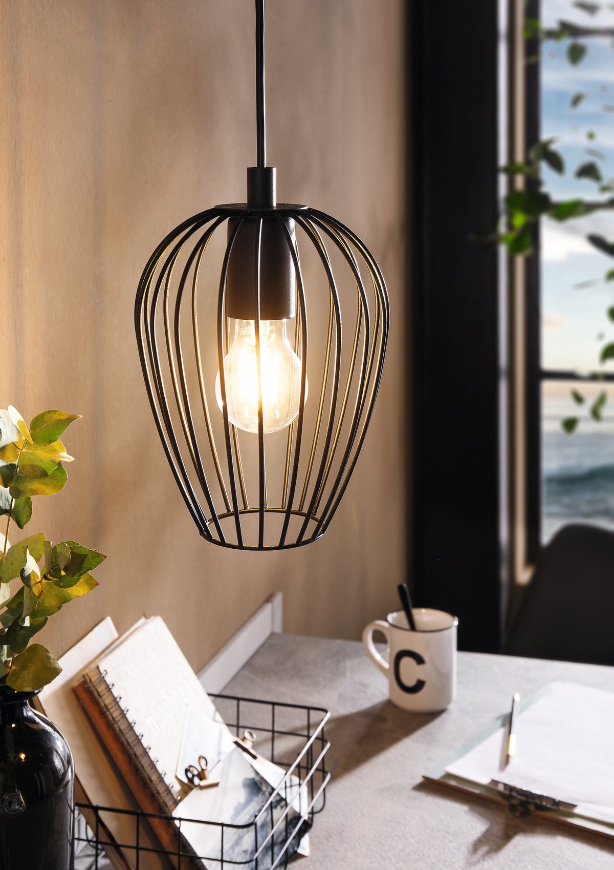 Mise en ambiance au-dessus d'un bureau, suspension noire allumée, plante et mug visibles.