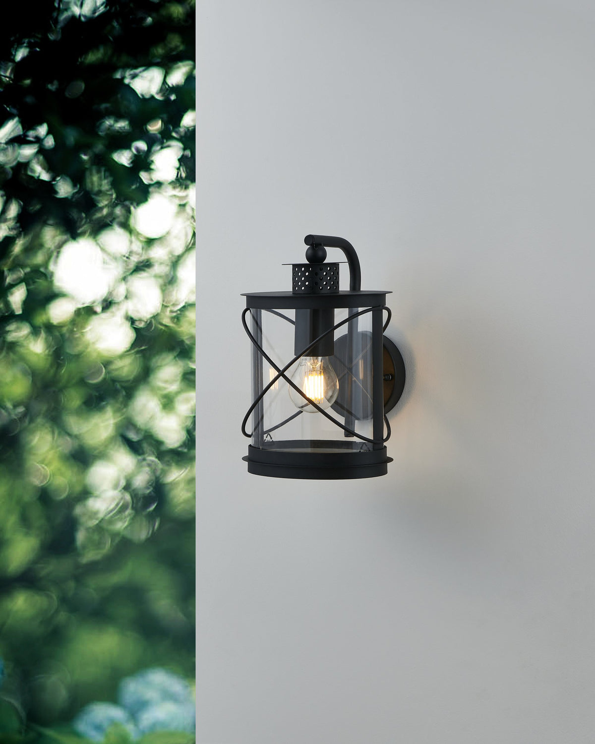 Mise en ambiance extérieure sur mur blanc, luminaire allumé près du feuillage