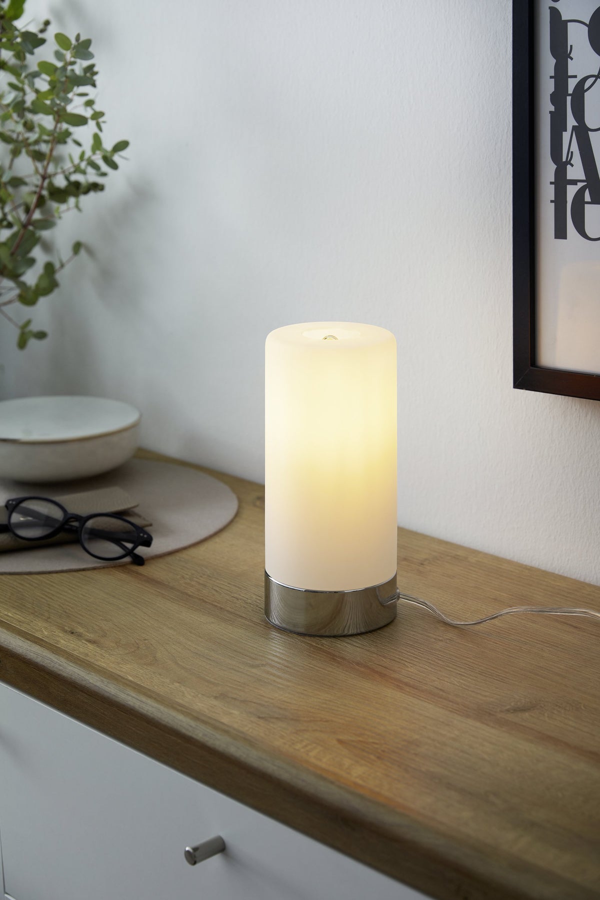 Mise en ambiance de la lampe allumée posée sur un meuble en bois clair, près d'une plante.
