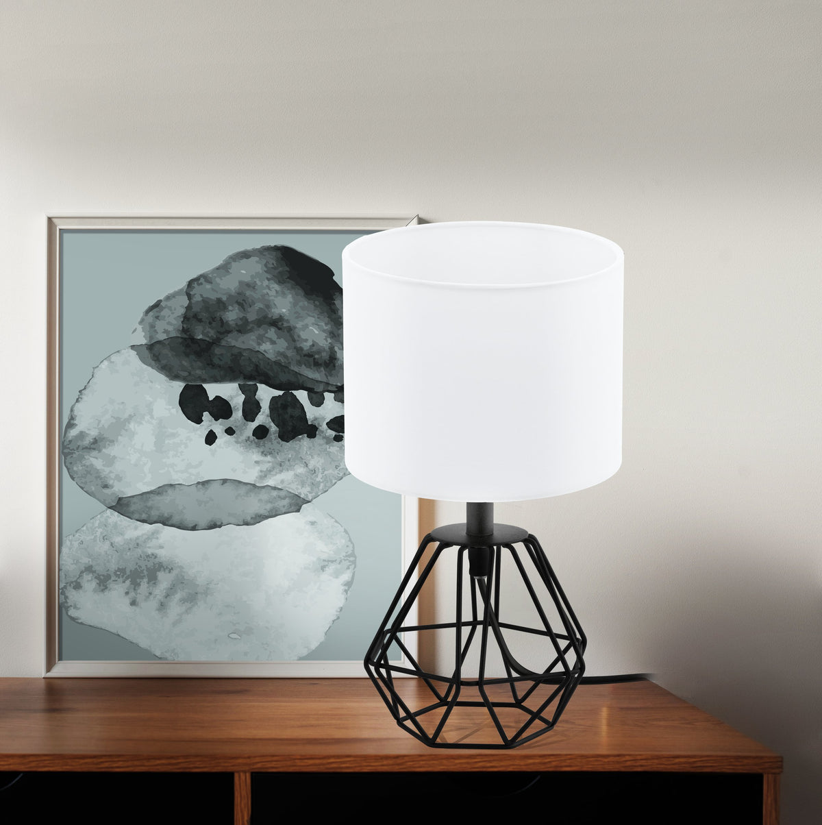Lampe posée sur une commode en bois, devant un tableau encadré, en situation