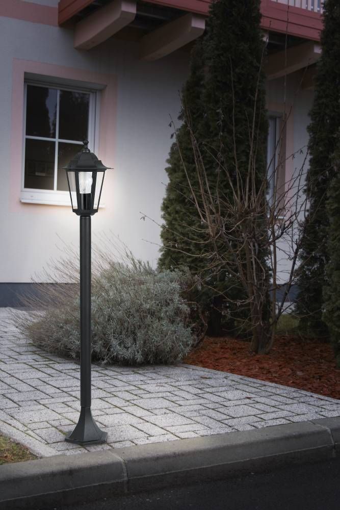 Lampadaire extérieur allumé près d'une maison, installé sur des pavés, avec arbustes