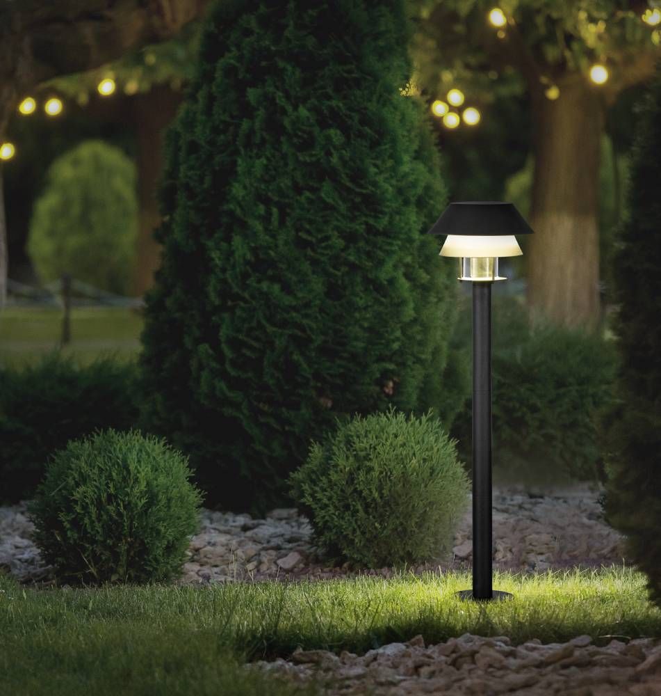 Lampadaire éclairé dans un jardin de nuit, entouré de buissons et de graviers.