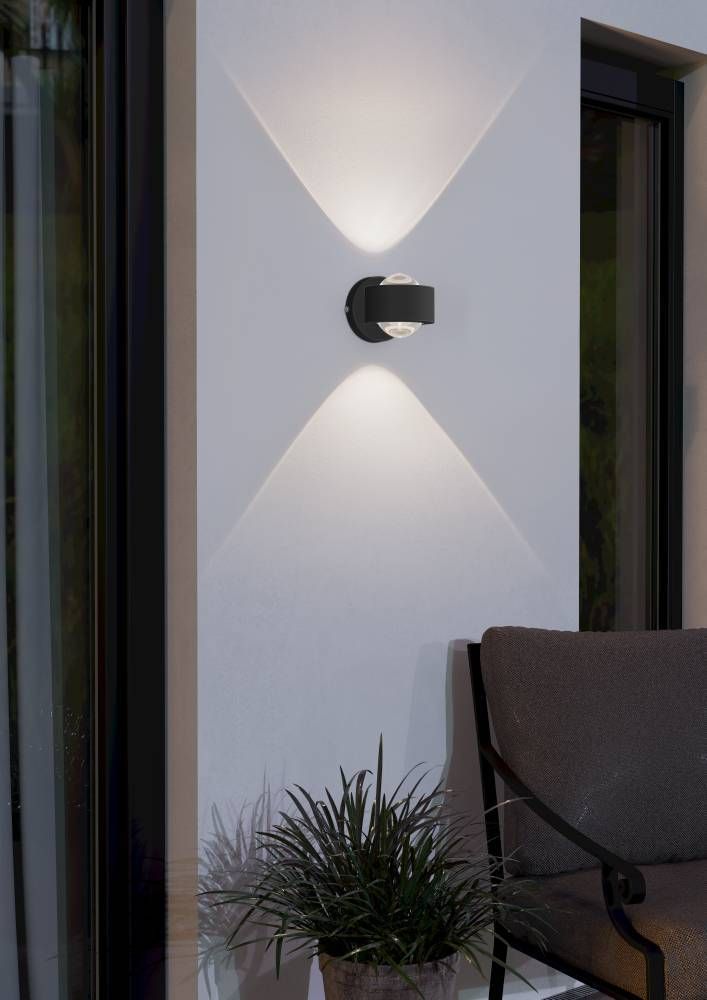 Mise en ambiance extérieure : applique murale noire éclairant un mur blanc, plante et fauteuil.