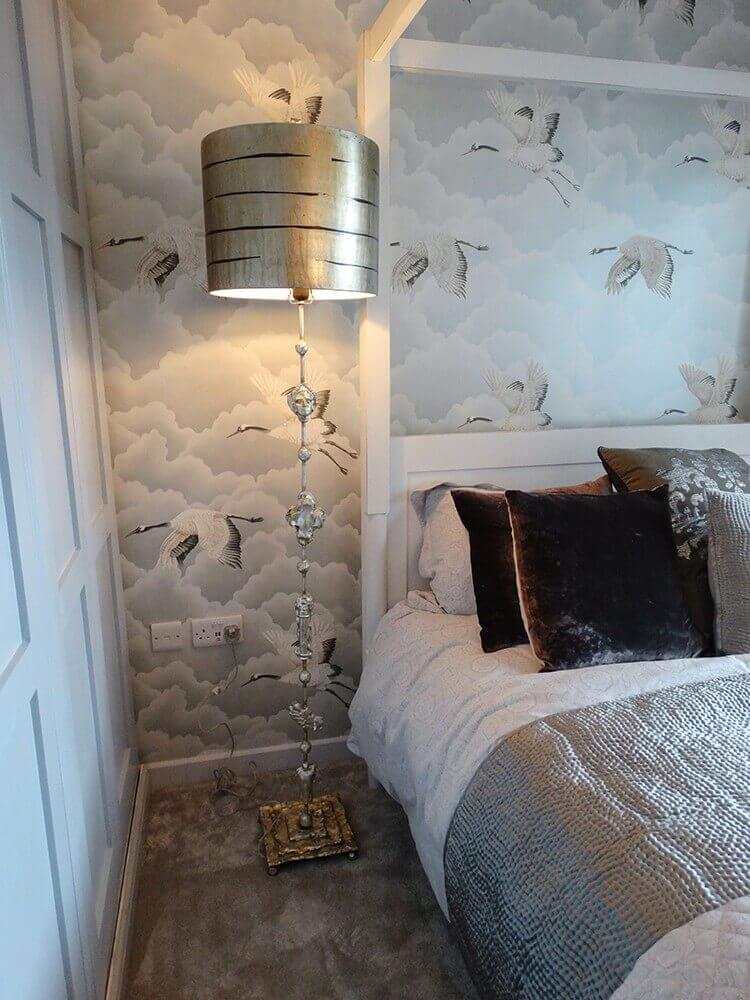 Lampadaire doré installé dans une chambre près du lit, papier peint à grues et nuages.
