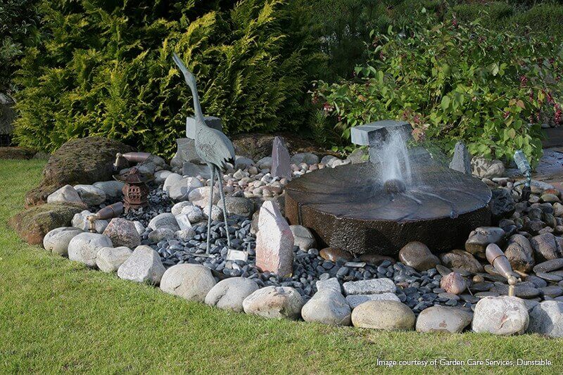 Mise en ambiance : jardin paysager, fontaine, rochers, statue de héron, sans luminaire visible.