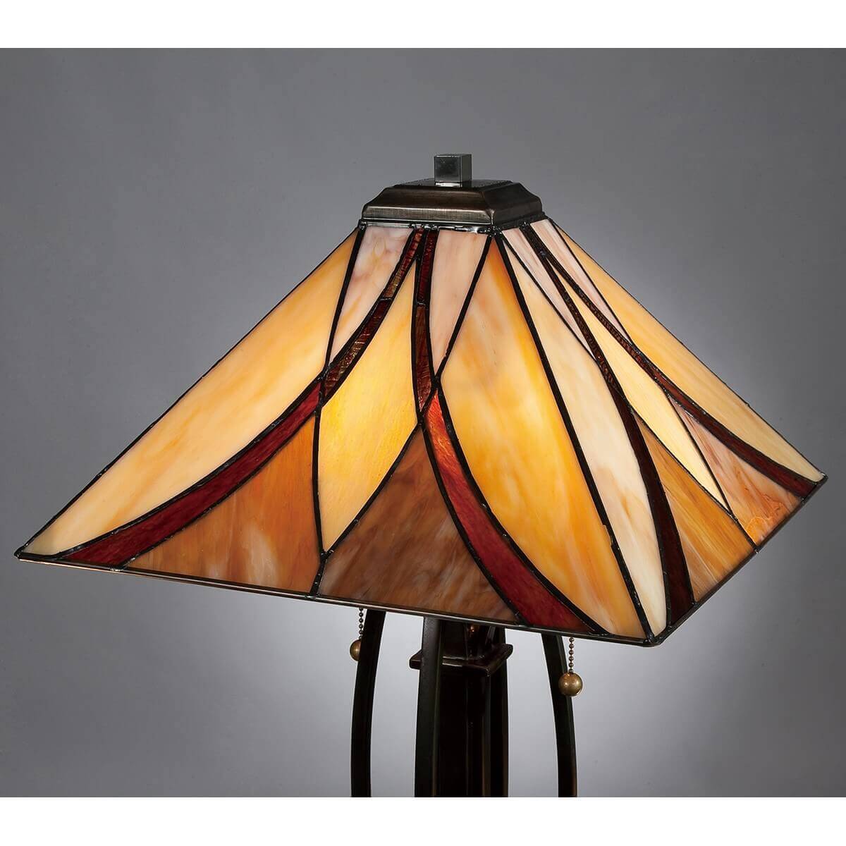 Gros plan sur l’abat-jour en verre Tiffany allumé, motifs géométriques rouge et ambre sur fond sombre.