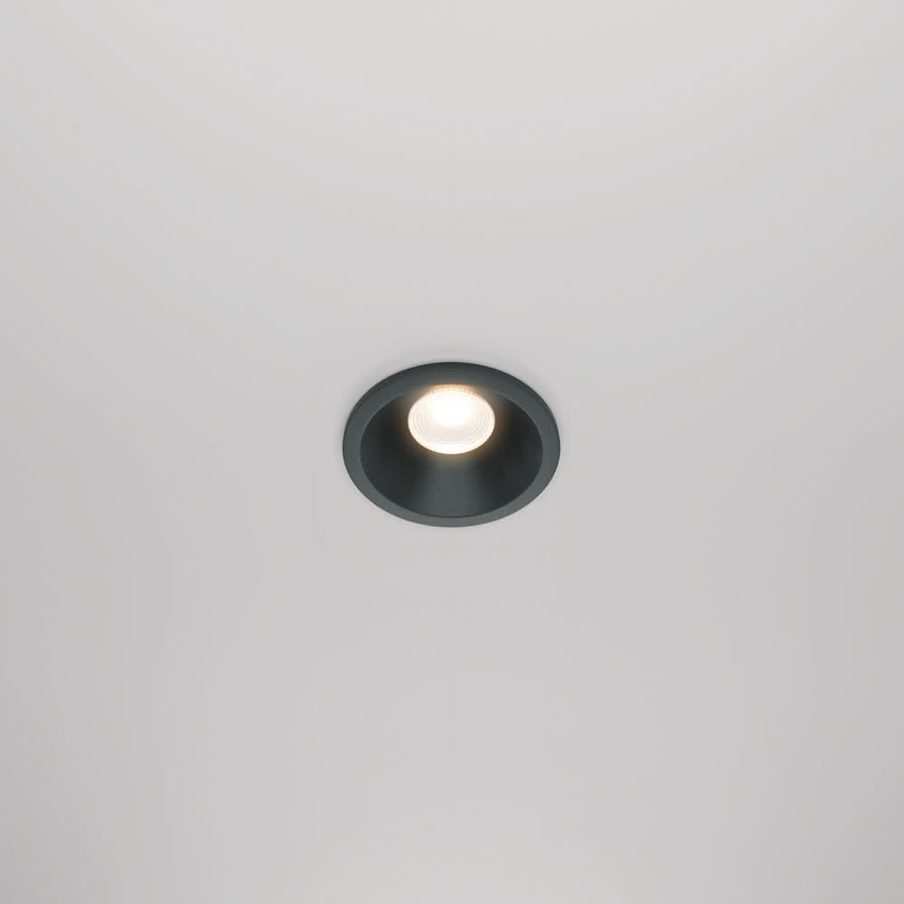 Spot encastré rond noir allumé, vu de face dans un plafond blanc