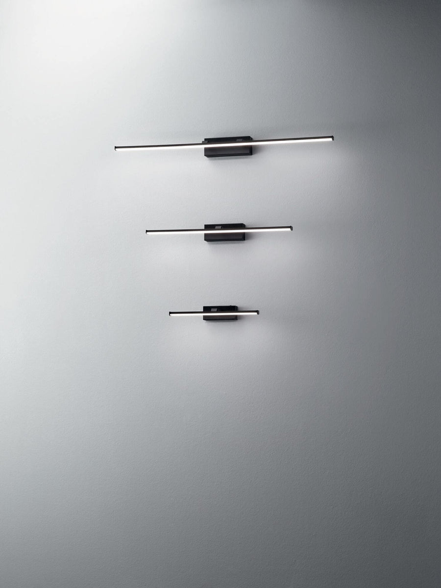 Trois appliques murales noires de tailles différentes alignées verticalement sur un mur gris