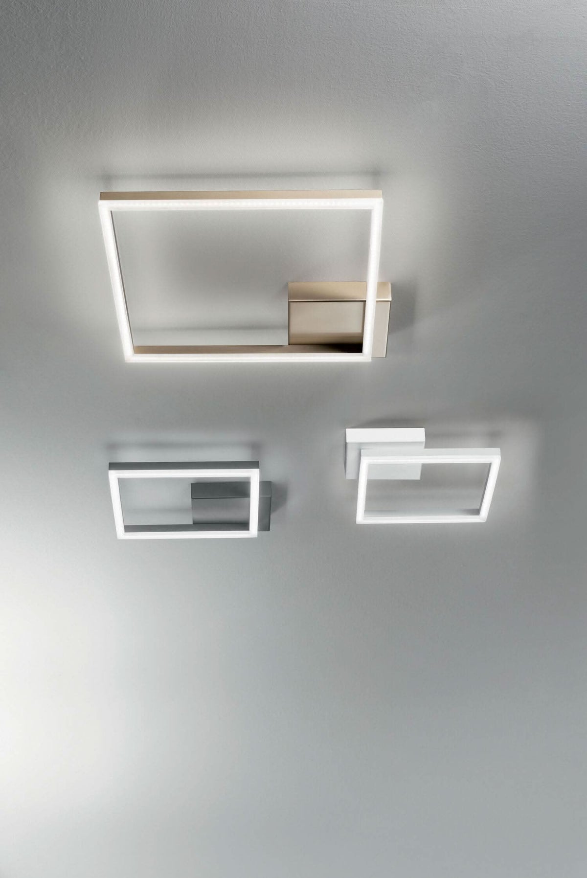 Trois plafonniers carrés fixés au plafond gris, finitions dorée, anthracite et blanche