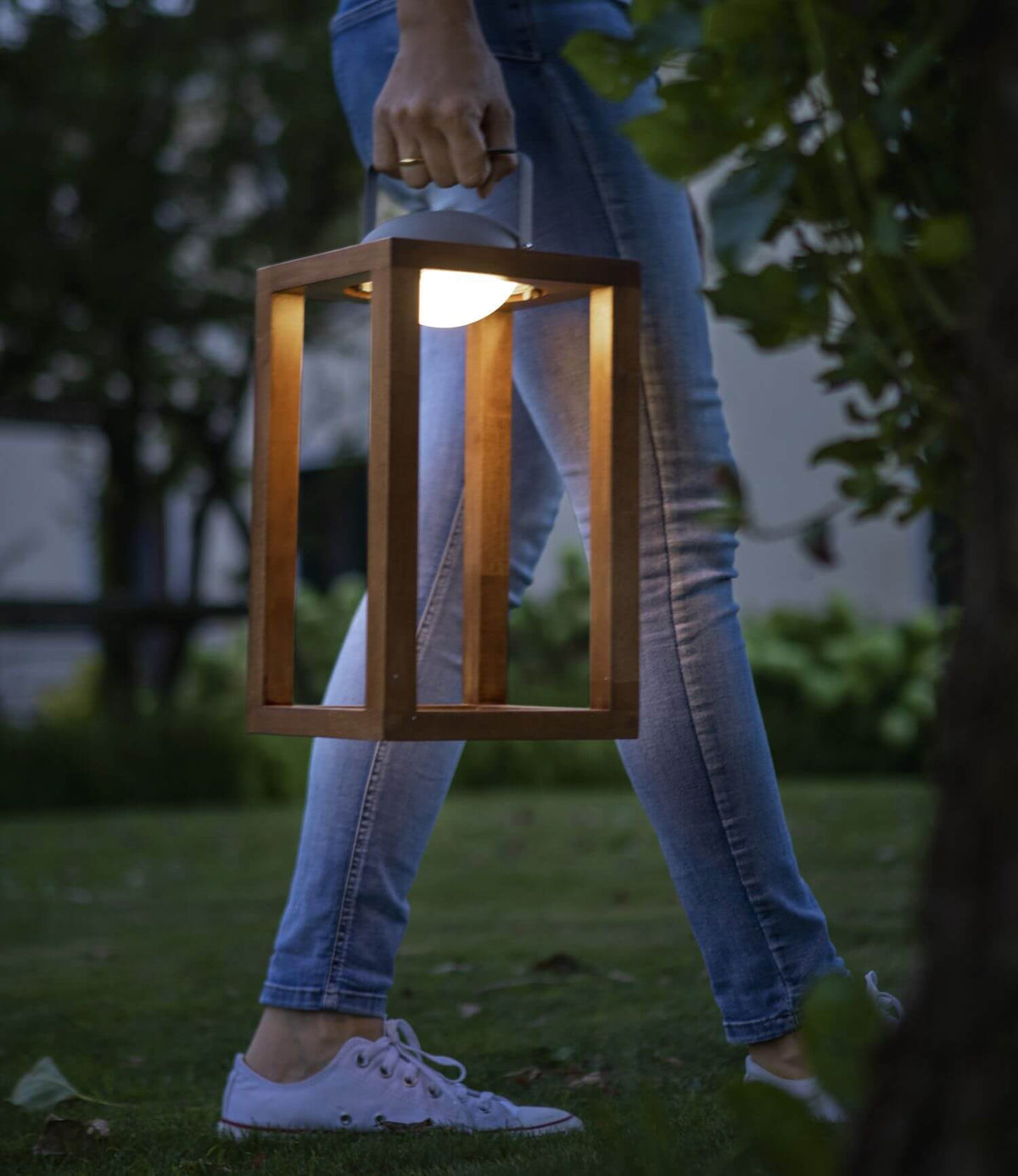 Personne portant la lanterne par sa poignée dans un jardin au crépuscule.