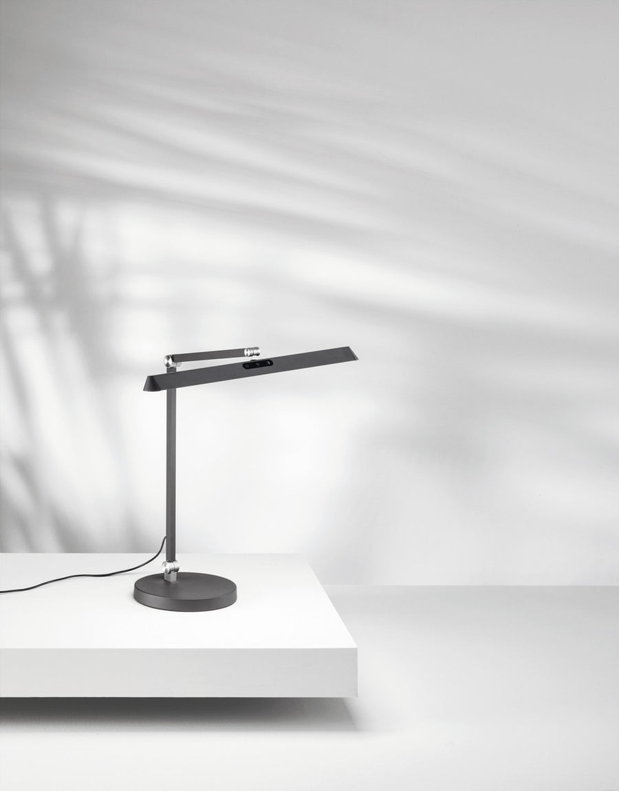 Lampe de bureau anthracite sur surface blanche, ombres portées et câble visible.