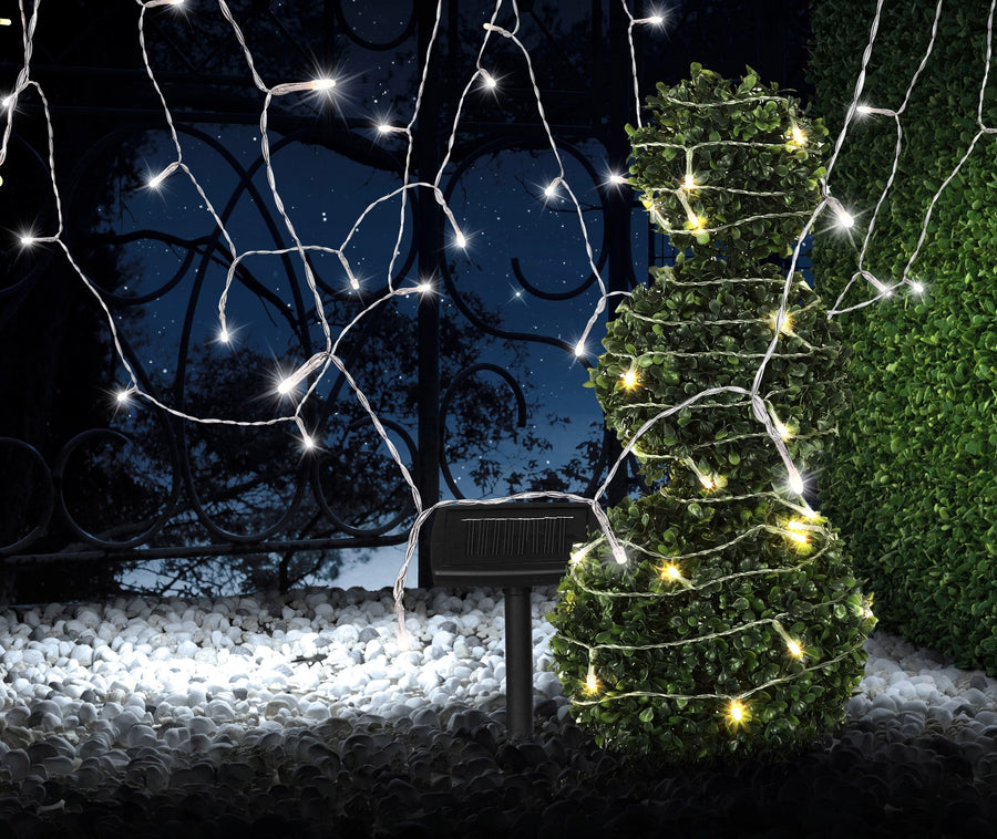 Scène nocturne : guirlande solaire allumée autour d'un topiaire, piquet solaire sur graviers
