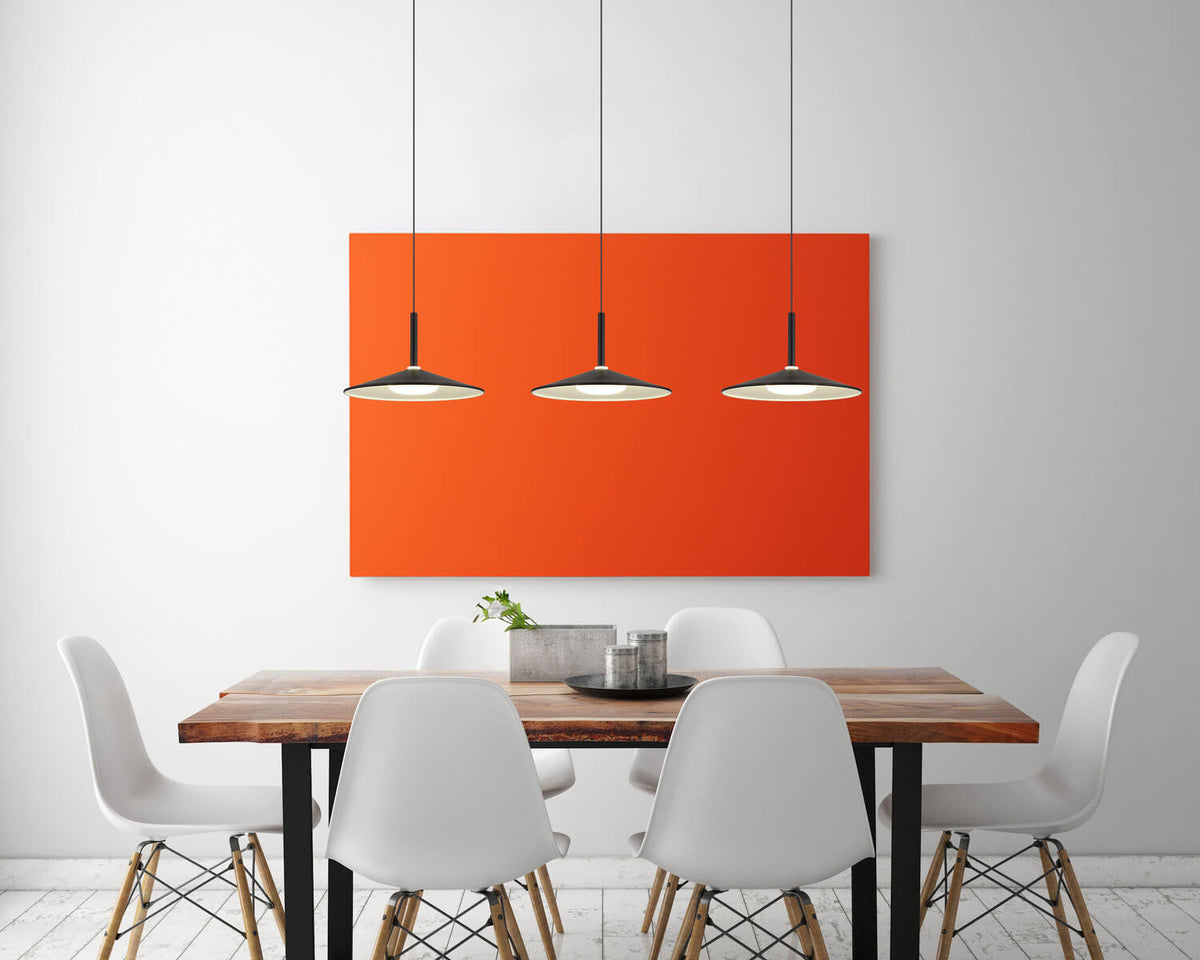 Mise en ambiance au-dessus d'une table à manger, trois suspensions noires alignées devant un tableau orange.