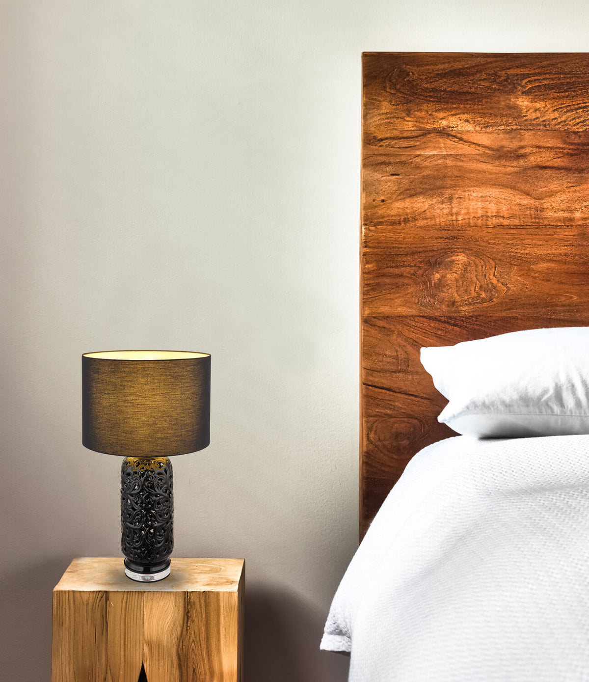 Mise en ambiance : lampe allumée sur une table de chevet en bois, tête de lit foncée