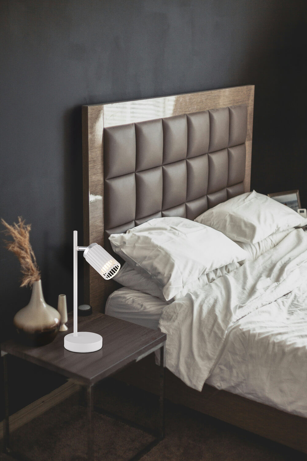 Mise en ambiance dans une chambre sombre, lampe posée sur table de chevet près du lit.