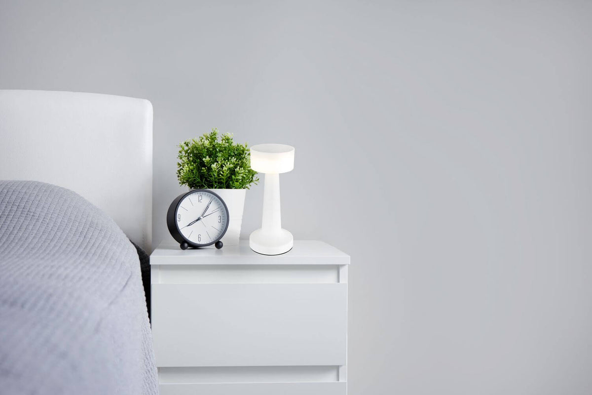Lampe blanche posée sur une table de chevet, près d'un réveil et d'une plante.