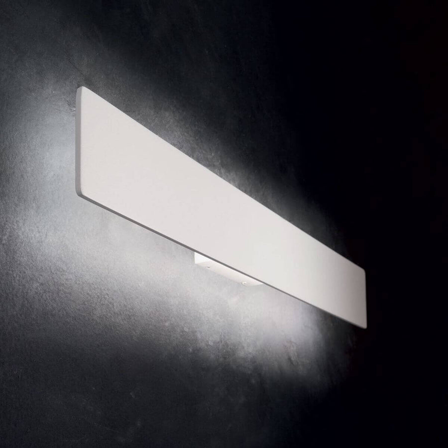 Applique murale blanche allumée sur un mur sombre, éclairage haut et bas.