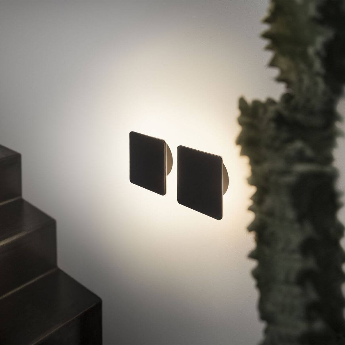 Deux appliques carrées noires vues de profil, halo lumineux diffus sur le mur