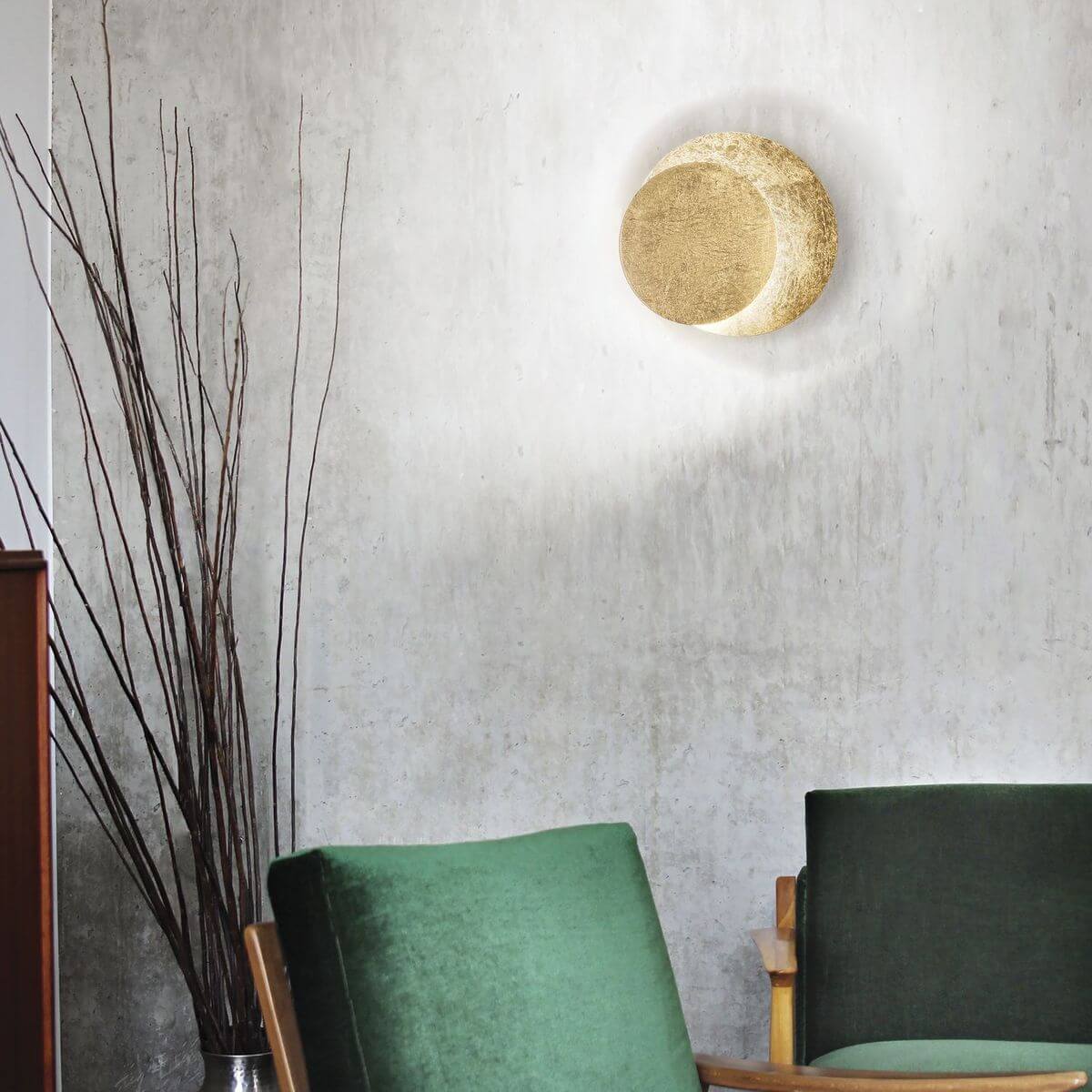 Luminaire doré installé sur un mur béton gris, ambiance salon avec fauteuils verts