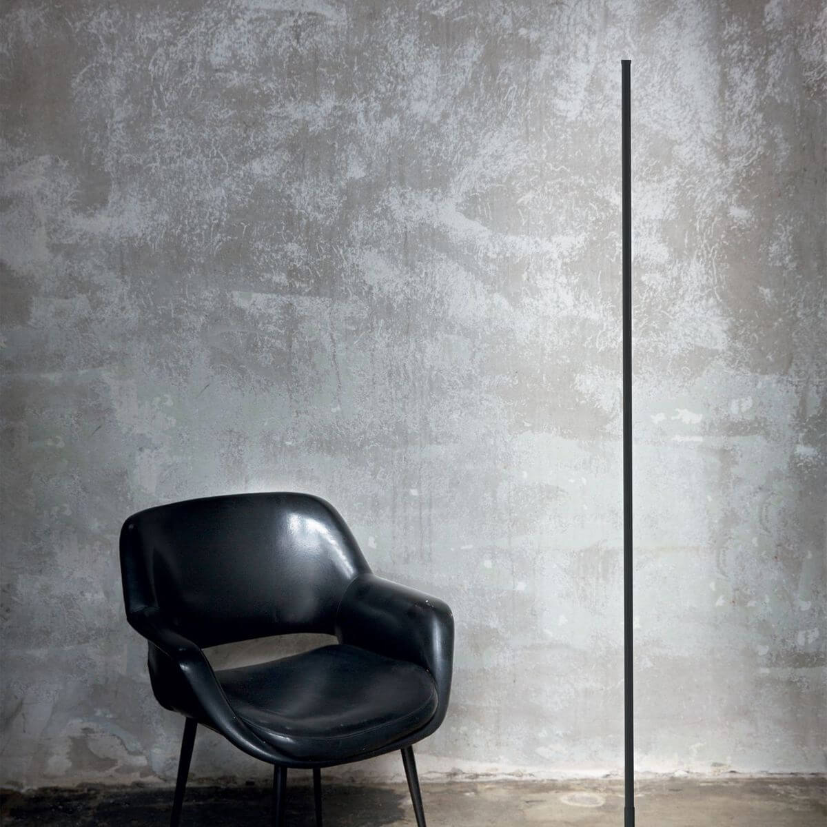 Mise en ambiance du lampadaire noir vertical près d'un fauteuil noir devant mur béton.