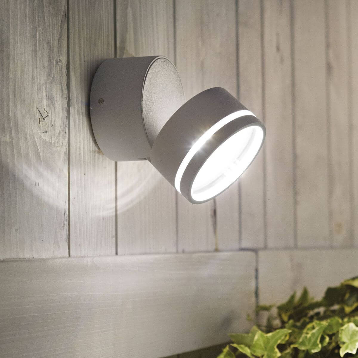 Applique allumée fixée sur bardage bois blanc, halo lumineux et plantes vertes en ambiance extérieure.