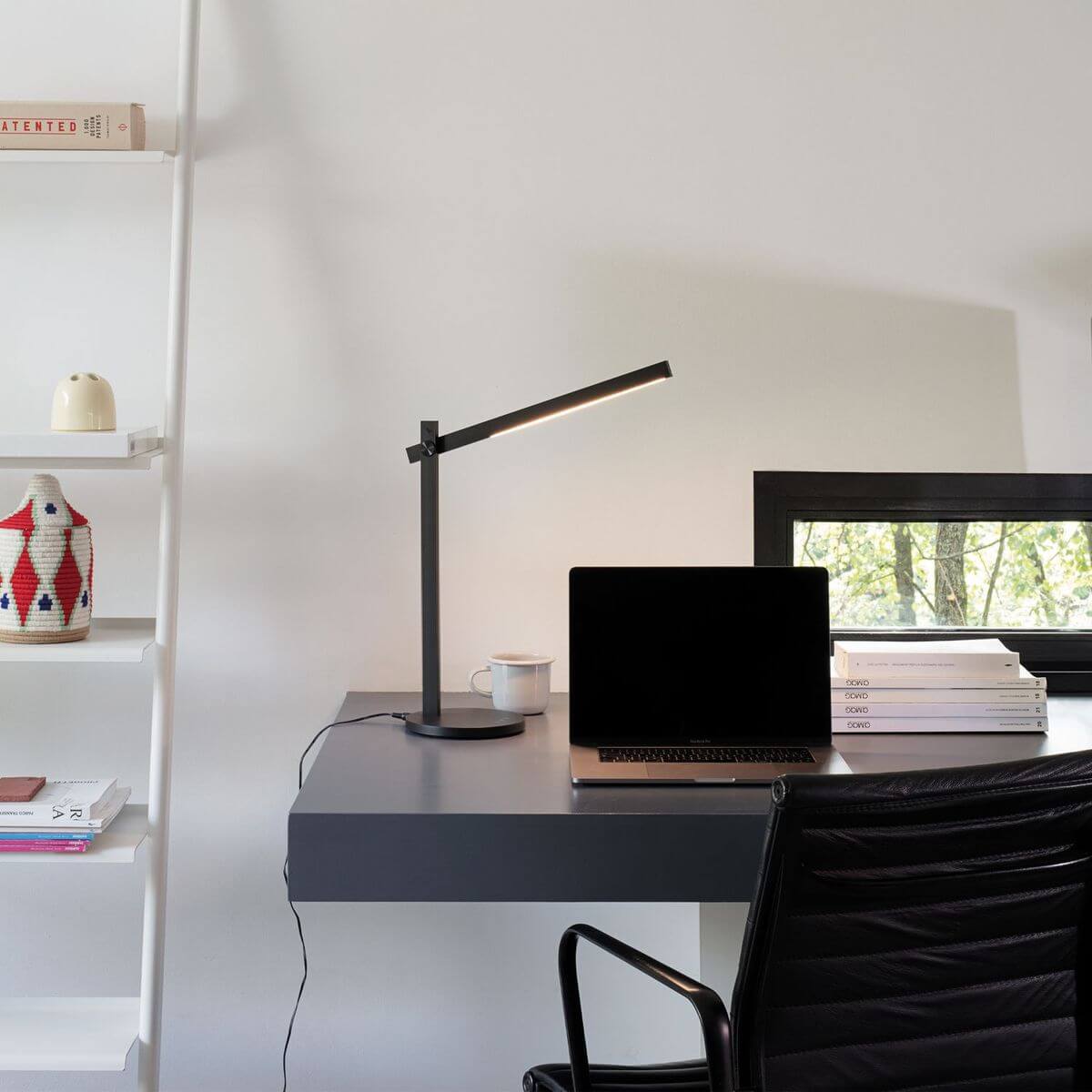 Lampe noire posée sur bureau gris, près d'un ordinateur et d'une fenêtre.
