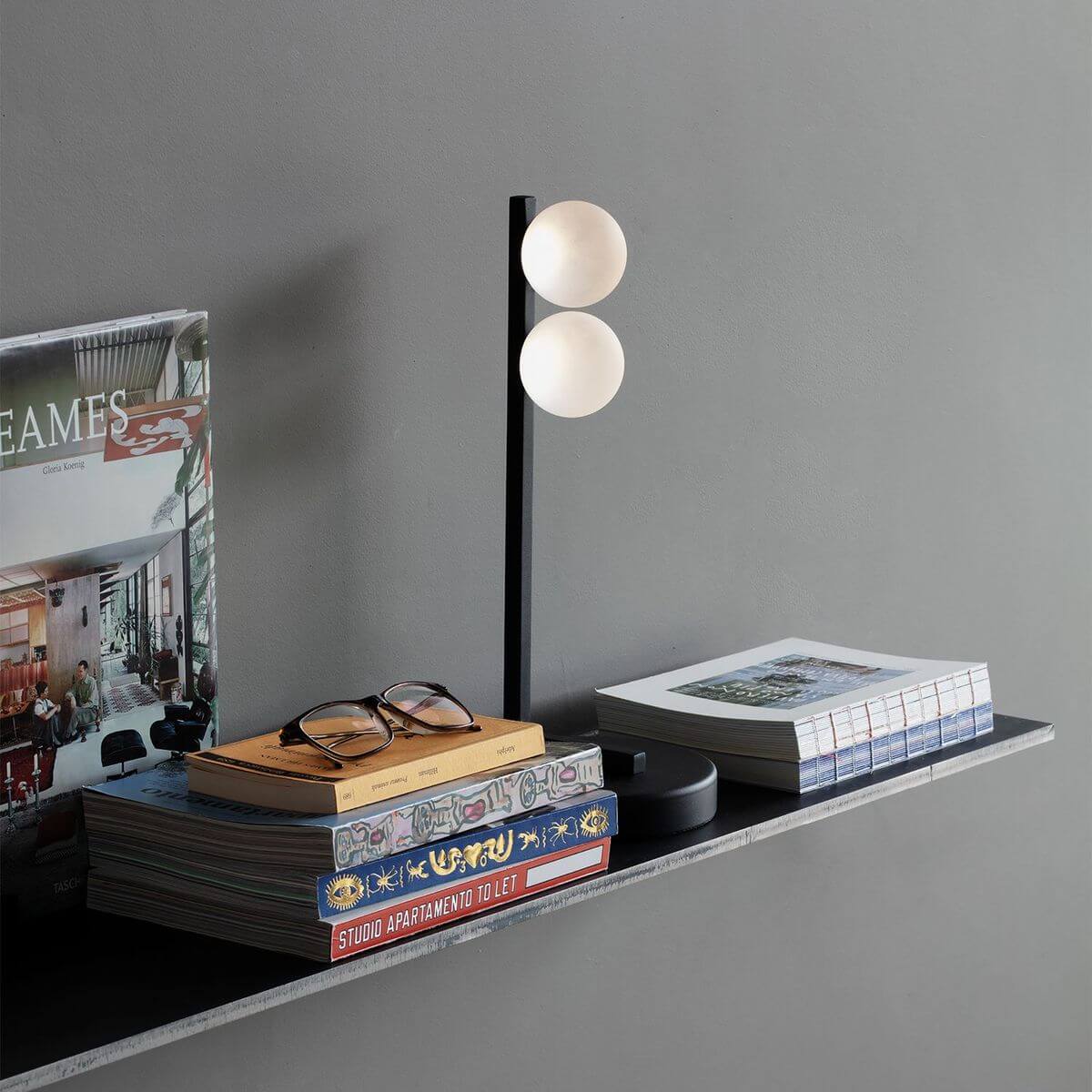 Mise en ambiance sur une étagère noire, livres, lunettes et lampe allumée.
