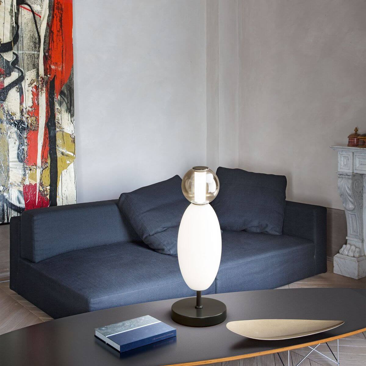 Mise en ambiance : lampe posée sur table basse devant canapé gris et tableau coloré.