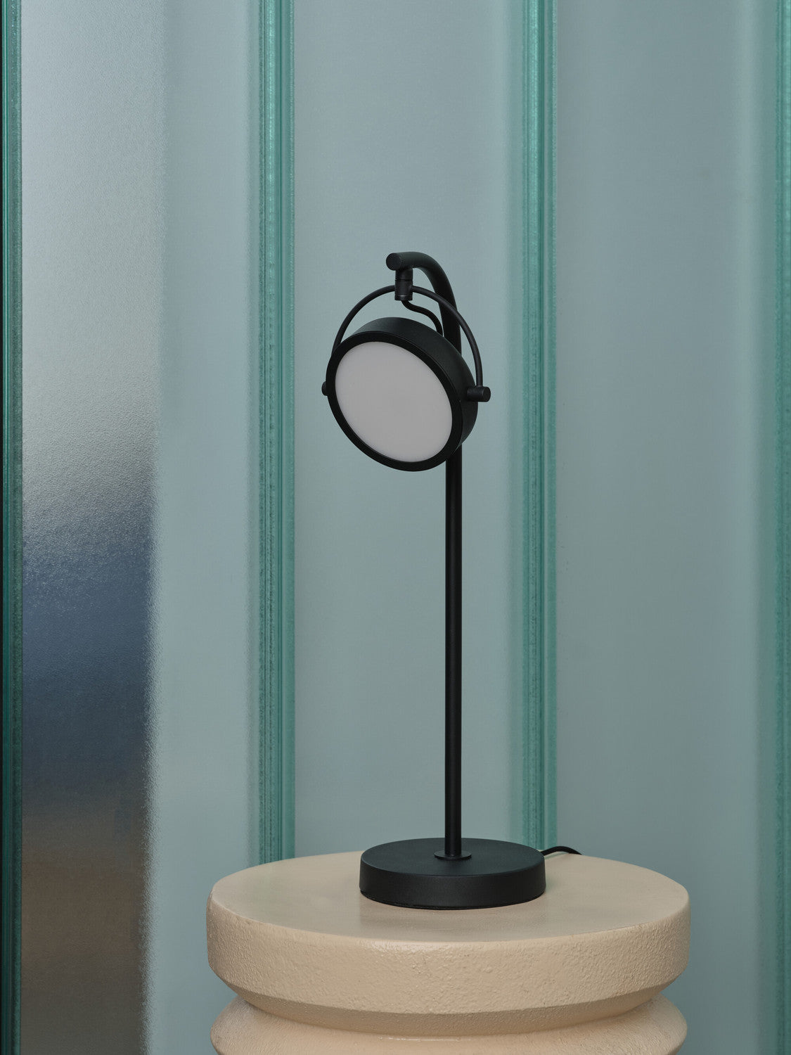 Lampe noire vue de face sur socle beige, abat-jour rond et mur rayé.