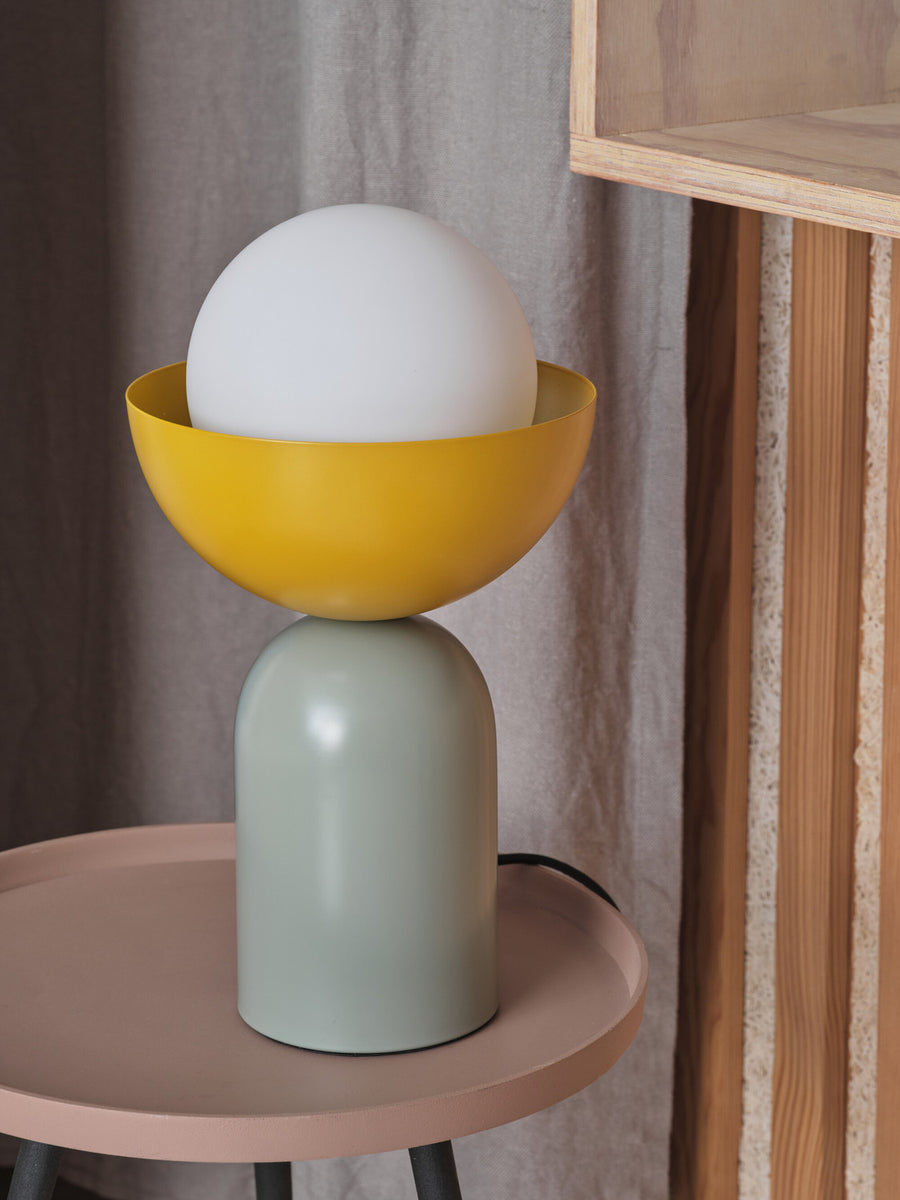 Gros plan du luminaire éteint posé sur une table, bol jaune et base verte.