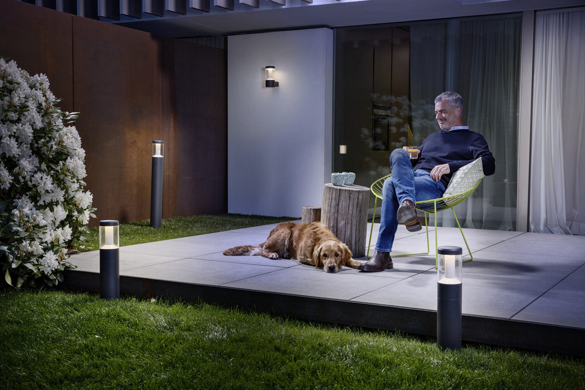 Ambiance nocturne sur une terrasse : homme assis, chien couché et luminaire mural allumé.