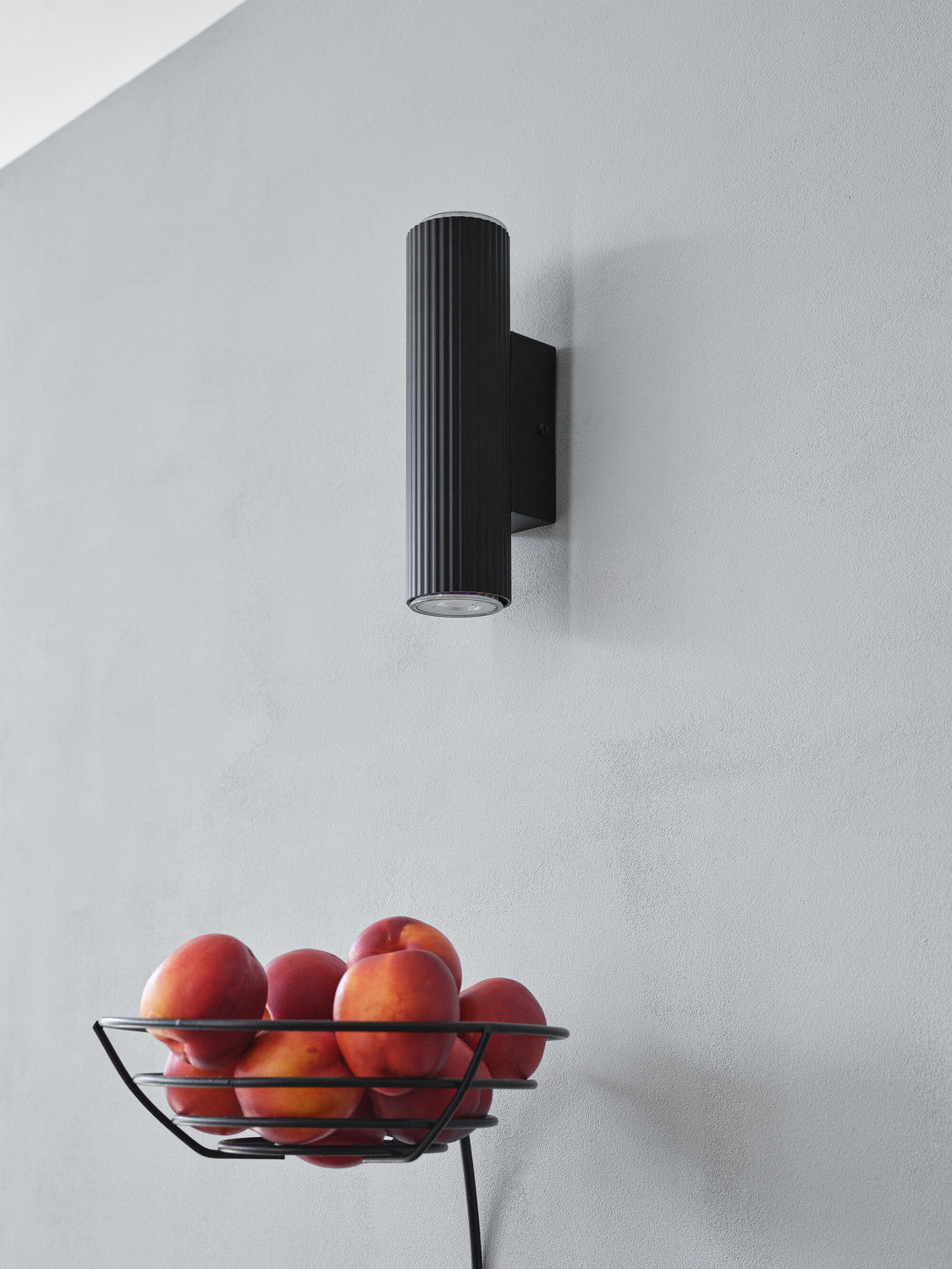 Applique cylindrique noire fixée sur mur gris, panier de fruits en dessous