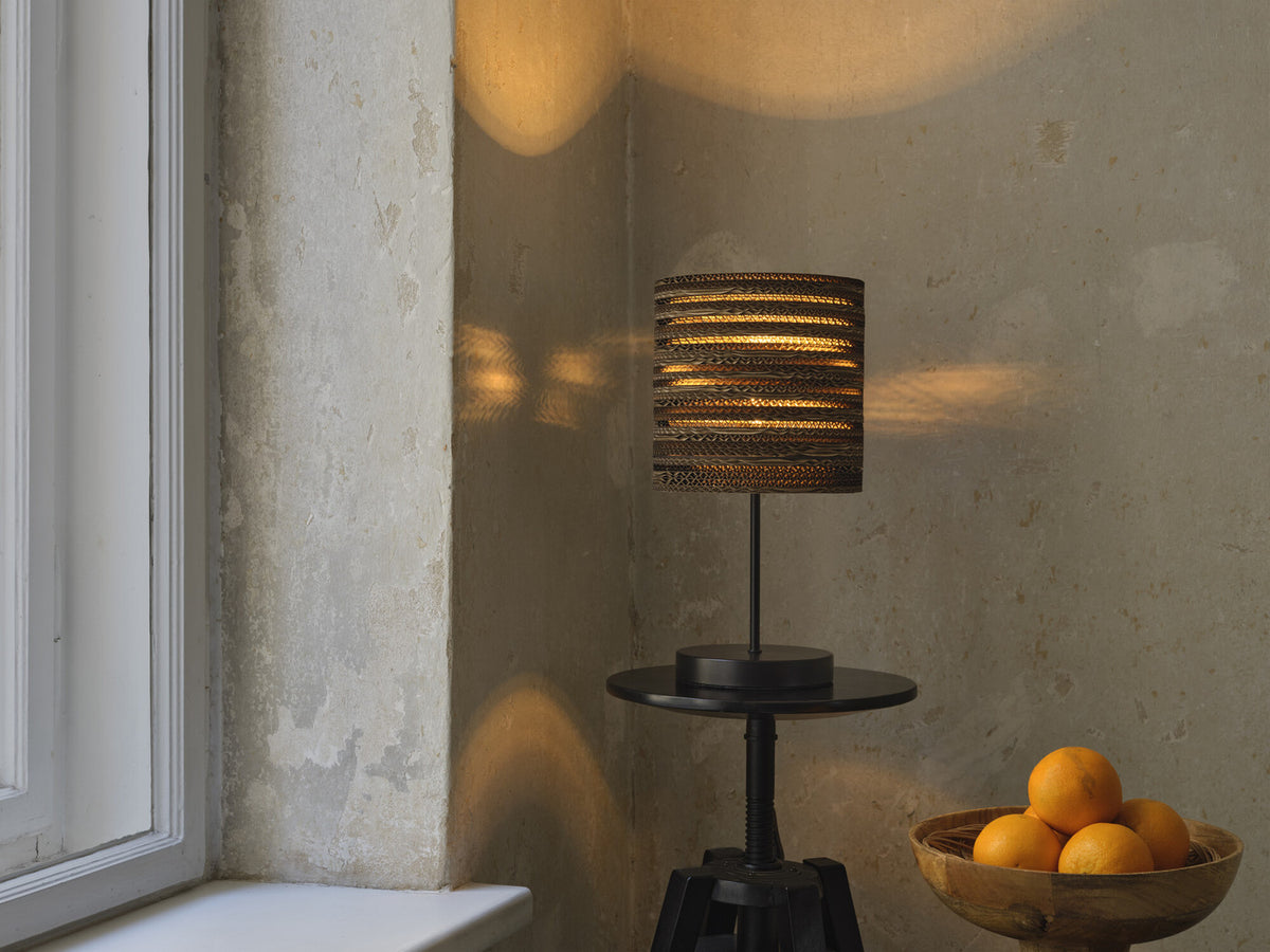 Lampe posée sur table noire près fenêtre, ombres dorées et bol d'oranges.