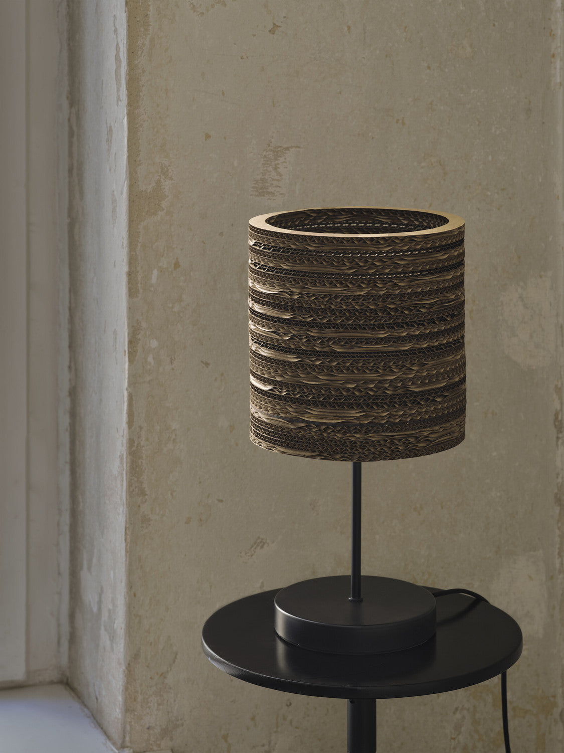 Lampe éteinte de face sur table noire, abat-jour carton et socle rond.
