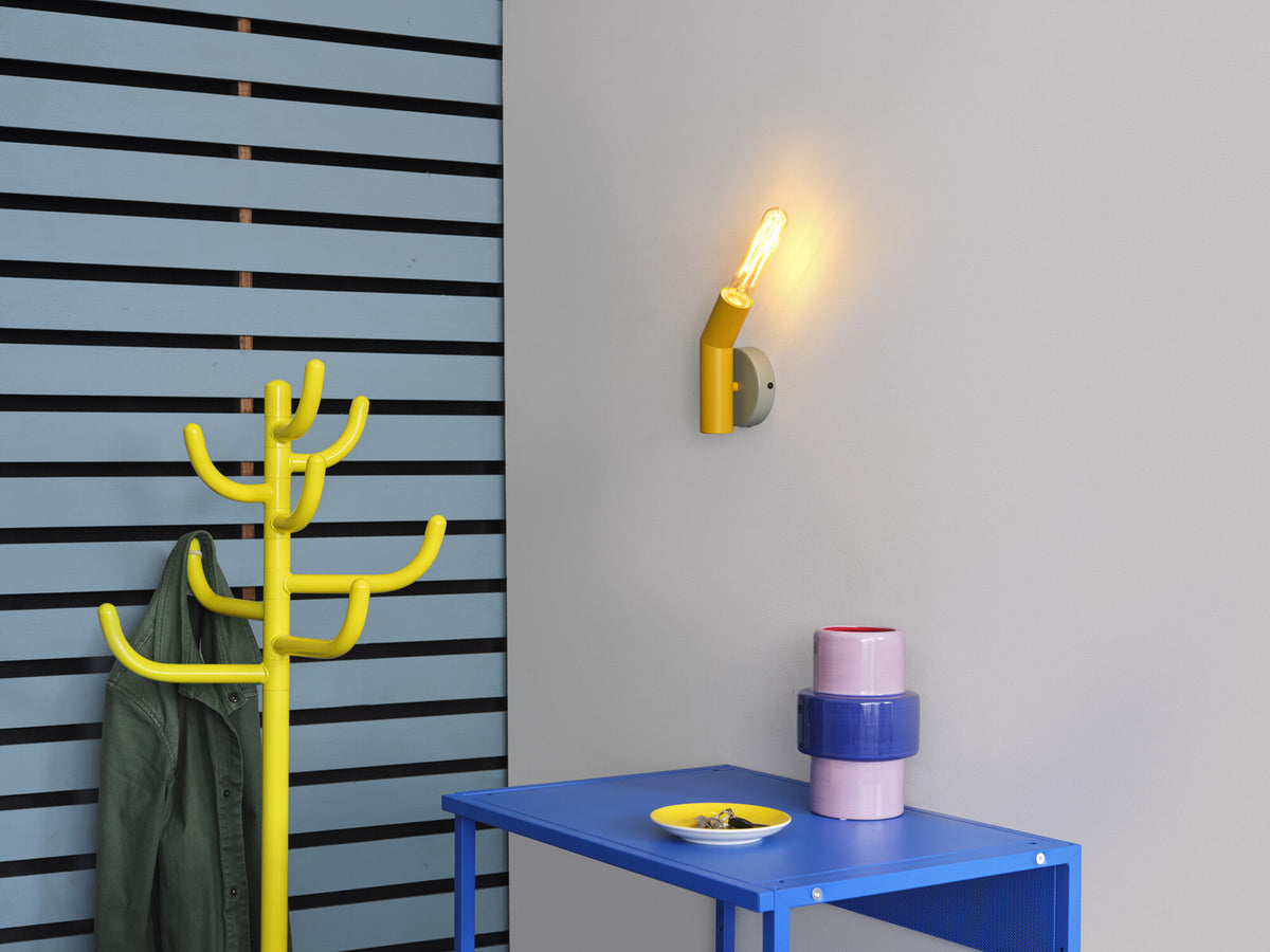Mise en ambiance sur mur gris clair, applique allumée près d’un portemanteau jaune.
