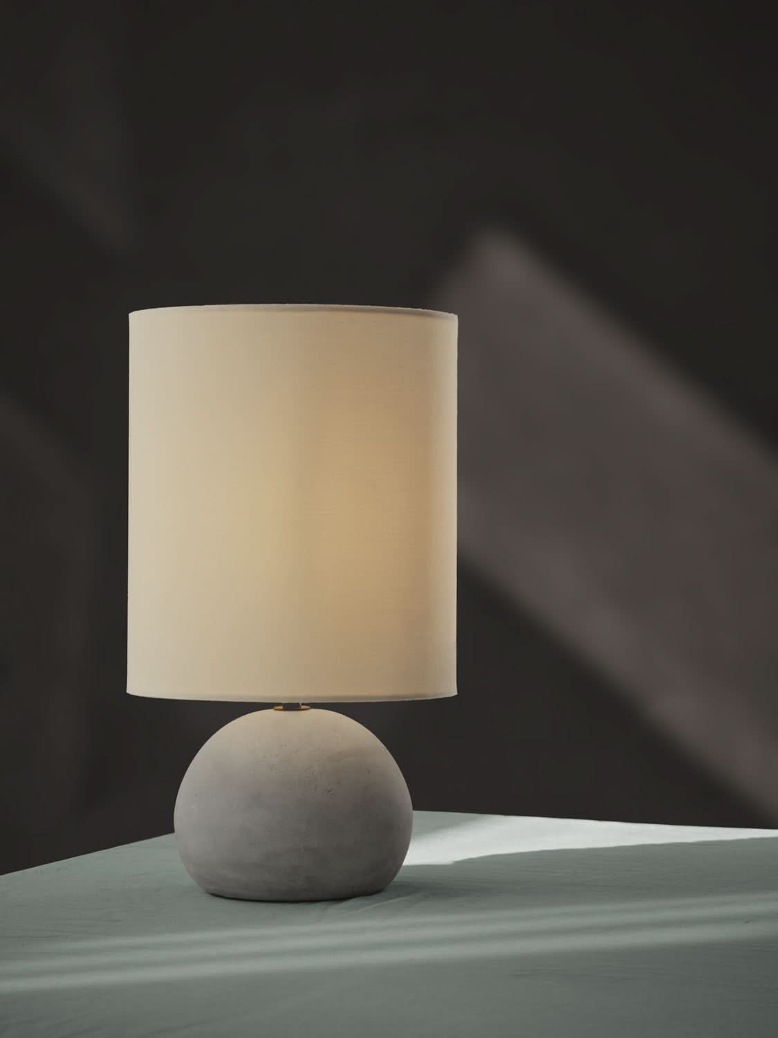 Vue de profil sur fond sombre, lampe allumée avec base sphérique en béton gris.