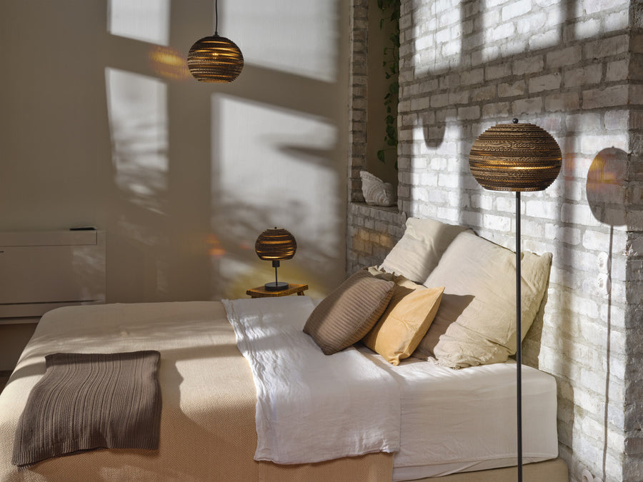 Suspension en carton éclairant une chambre avec lit, coussins et lampe sur pied.