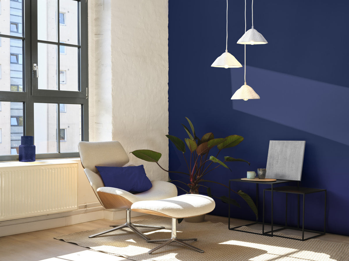 Suspension allumée au-dessus d’un fauteuil, dans un salon au mur bleu profond.