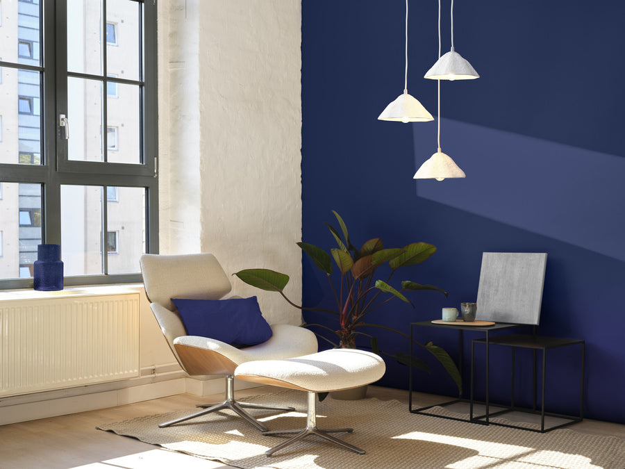 Suspension allumée au-dessus d’un fauteuil, dans un salon au mur bleu profond.