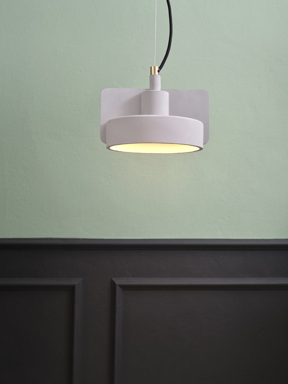 Suspension claire allumée, vue rapprochée sur mur vert et lambris noir, lumière chaude diffusée.