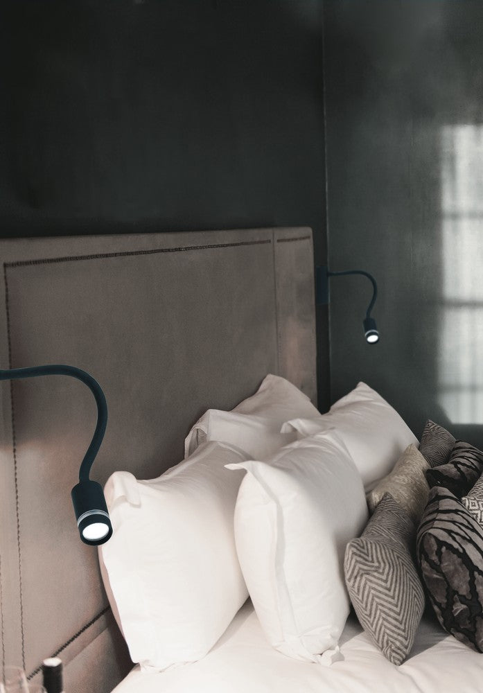 Mise en ambiance de deux appliques noires fixées au mur derrière une tête de lit claire.