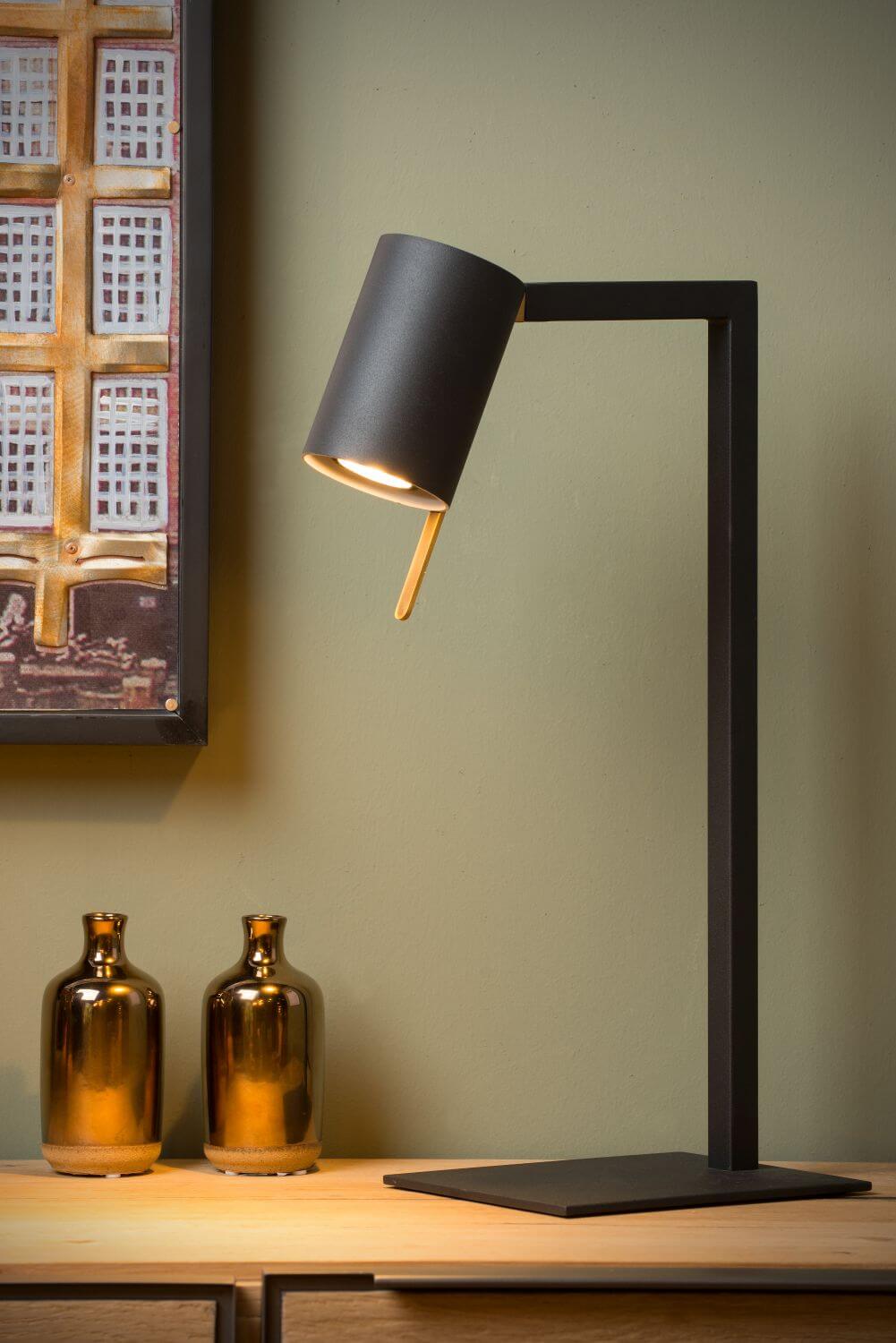 Lampe de bureau noire allumée sur un meuble en bois, près de deux petites bouteilles dorées.