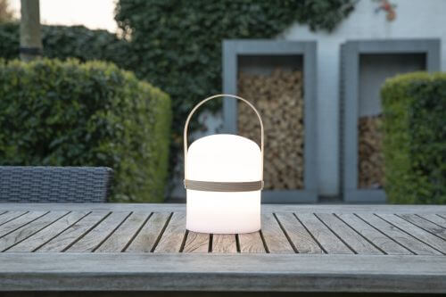 Lampe blanche allumée sur une table en bois, jardinières et haie taillée dans le jardin