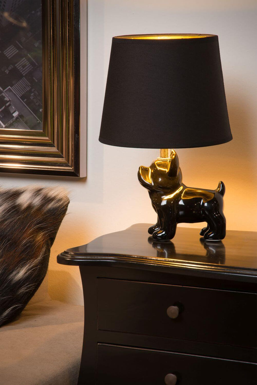 Lampe allumée posée sur une commode noire, cadre doré et coussin à motifs.