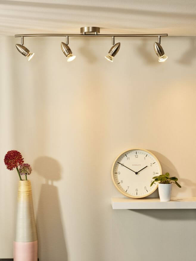 Plafonnier chromé allumé au plafond dans un intérieur avec horloge murale, vase et étagère.