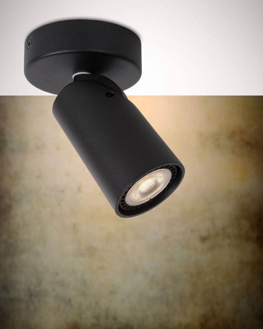 Spot cylindrique noir mat orientable vu de trois quarts sur fond texturé beige flou