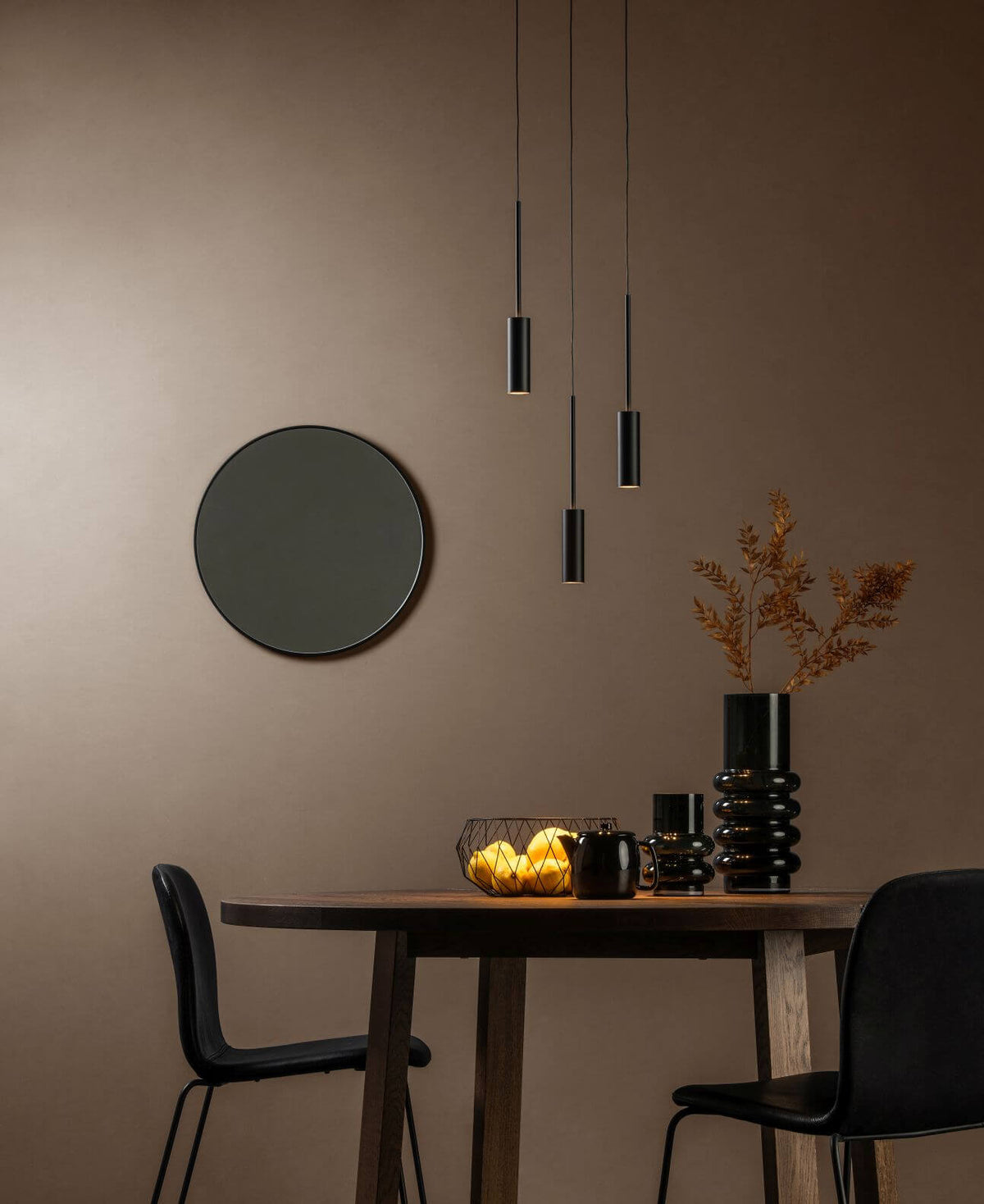 Suspension noire à trois tubes éclairant une table en bois dans un intérieur chaleureux.