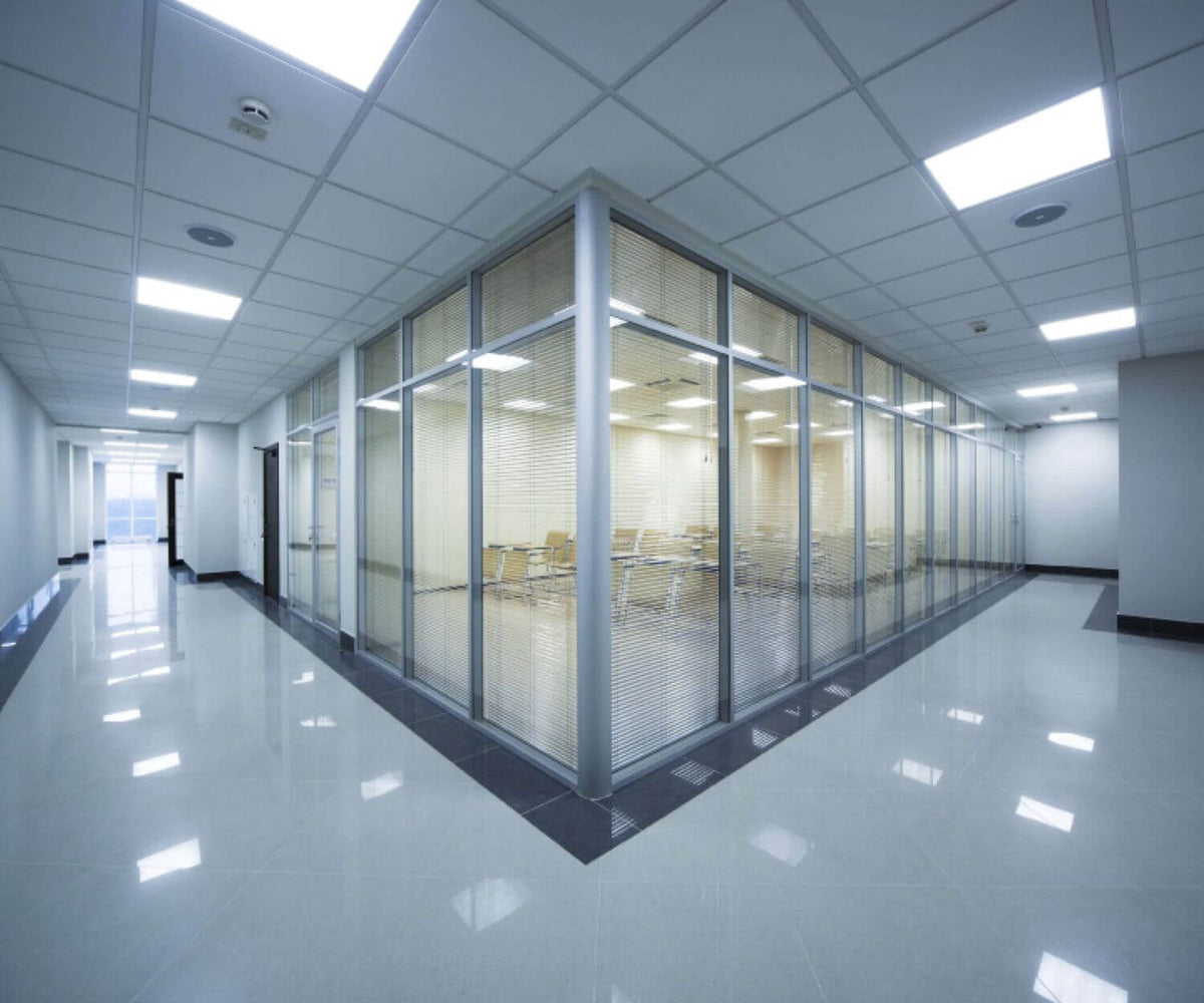 Long couloir de bureaux vitrés avec plafond technique et plusieurs luminaires encastrés allumés.