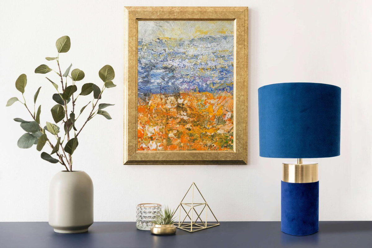 Lampe bleue et dorée devant un tableau encadré, avec vase et eucalyptus sur une console.